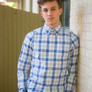 Young man in a plaid shirt leaning against a brick wall outdoors.