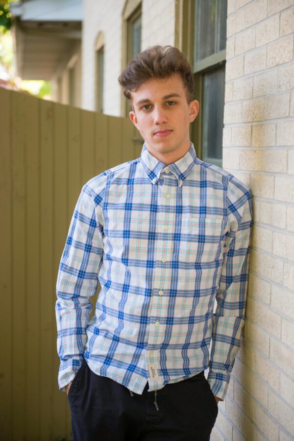 Young man in a plaid shirt leaning against a brick wall outdoors.